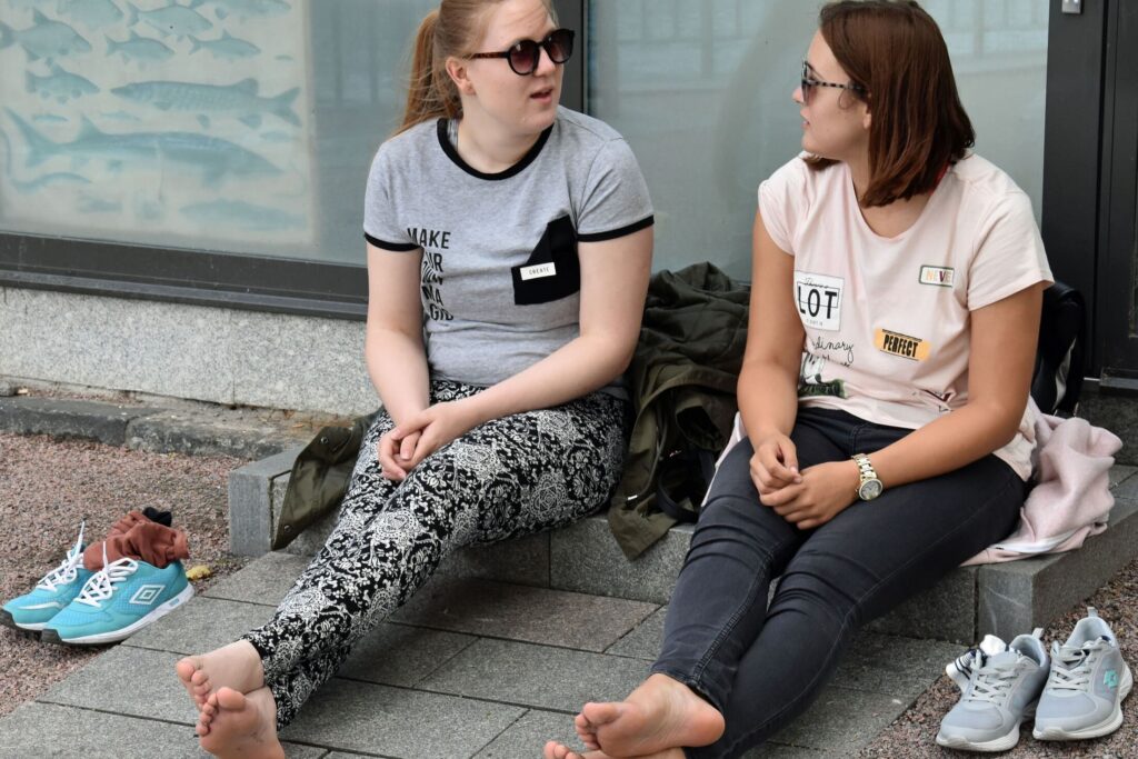 Dvije djevojke sa sunčanim naočalama, u kratkim rukavima i bez cipela i čarapa razgovaraju i sjede na betonskim stepenicama u Stockholmu, Švedska.
