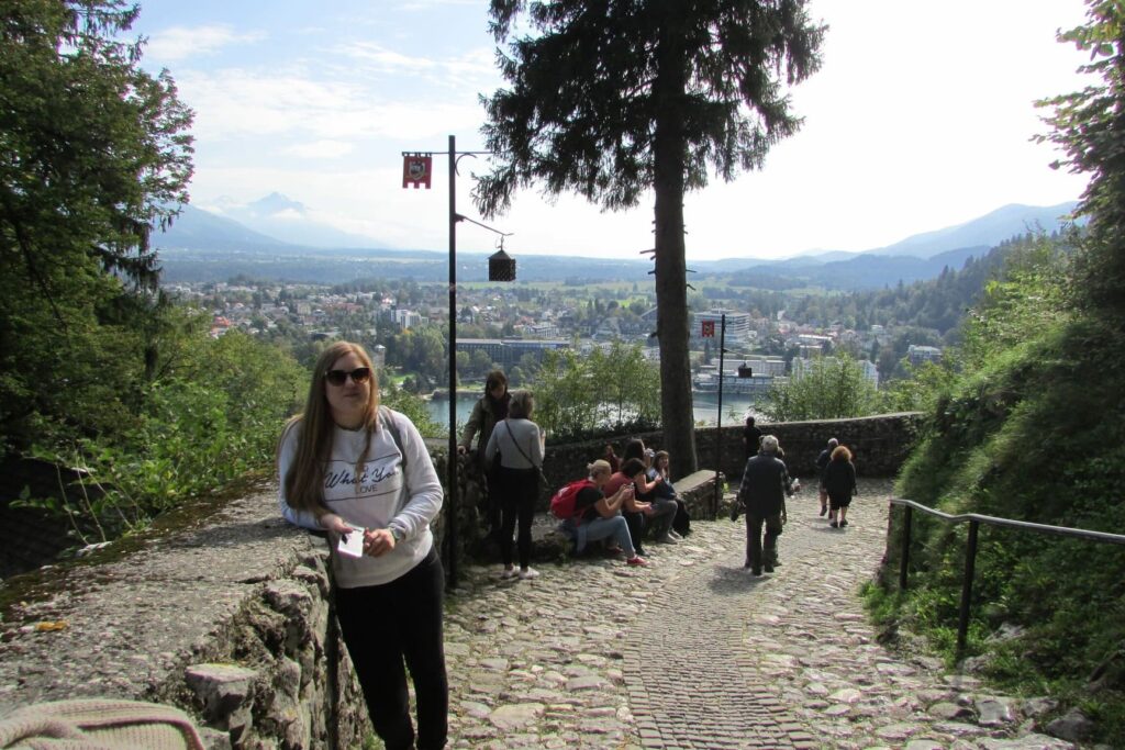 Djevojka s dugom riđom kosom, sunčanim naočalama u bijeloj majici i crnim hlačama naslonjena je na kameni zid s jezerom Bled i planinom Triglav u Sloveniji u pozadini.
