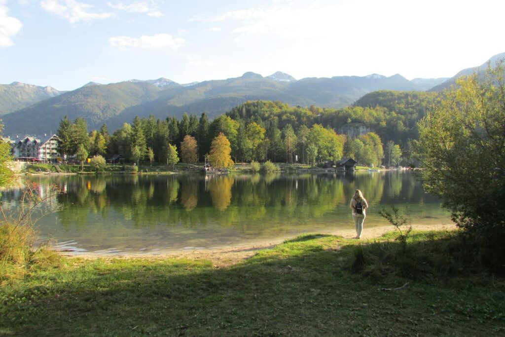 Djevojka okrenuta leđima stoji ispred jezera Bohinj u Sloveniji, koje je okruženo drvećem u jesenjskim bojama.