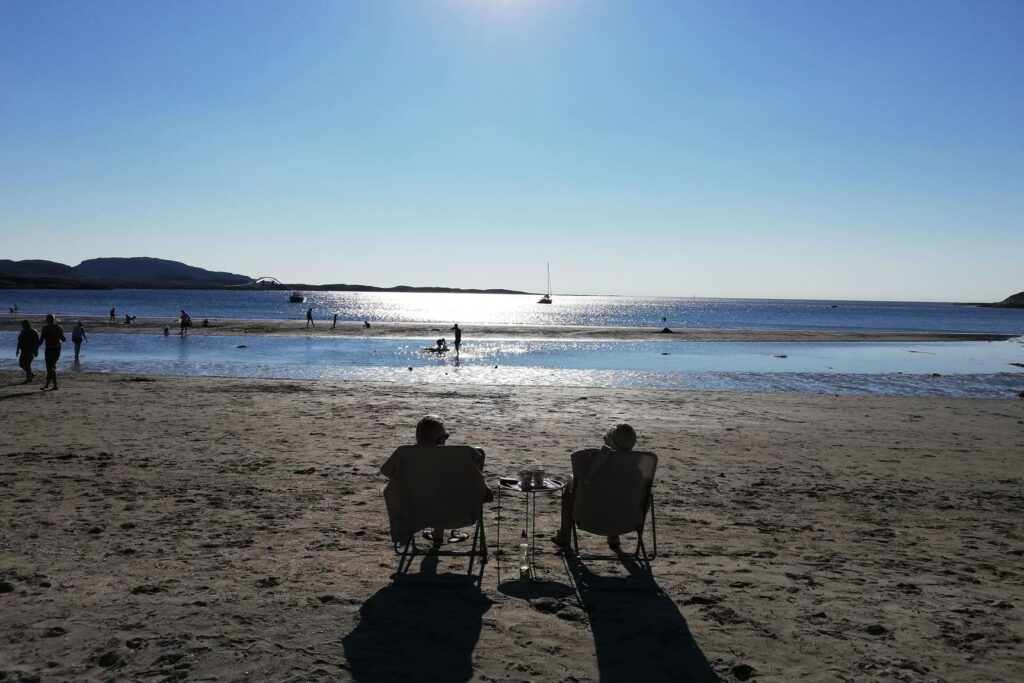 Dvoje ljudi fotografirani s leđa, sjede na sklopivim stolicama na pješčanoj plaži i gledaju prema Norveškom moru.