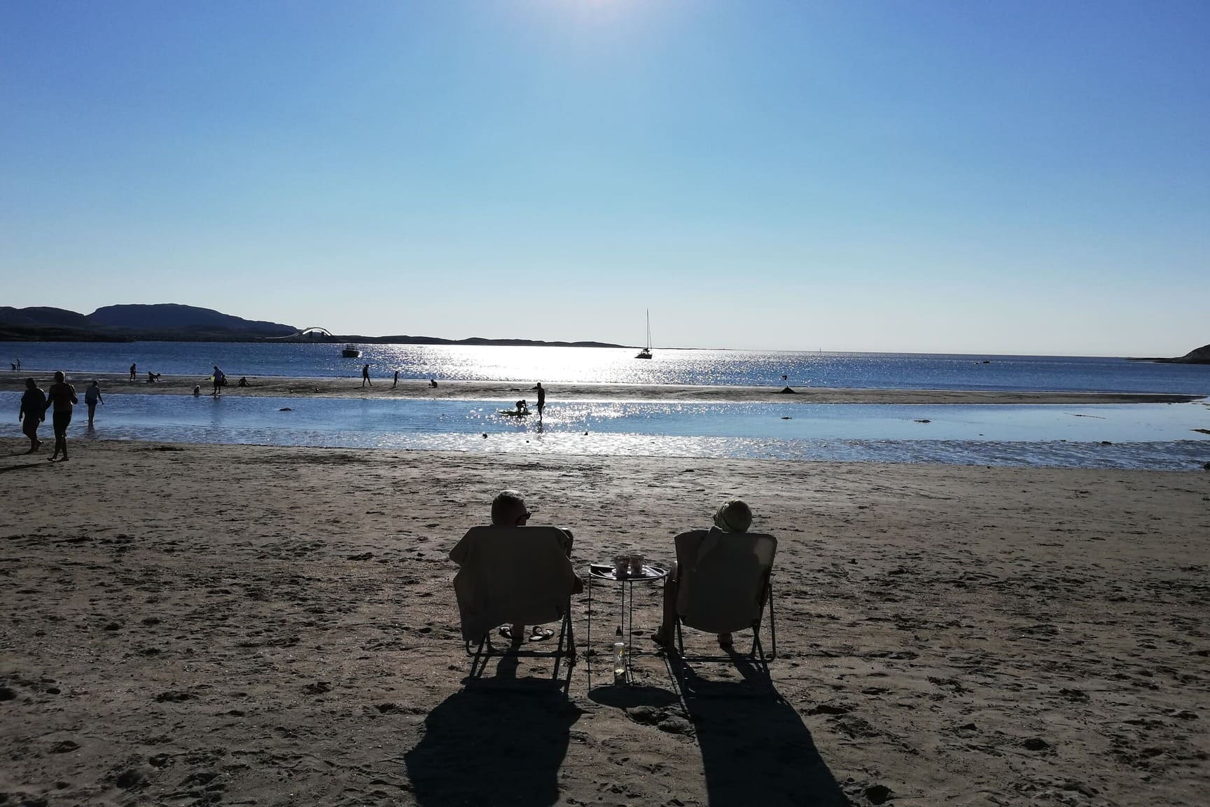 Dvoje ljudi fotografirani s leđa, sjede na sklopivim stolicama na pješčanoj plaži i gledaju prema Norveškom moru.
